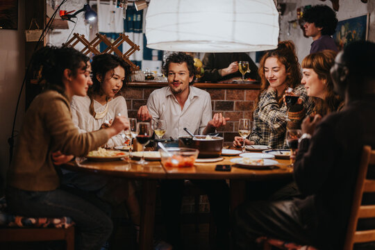 A group of people gather around a table enjoying food and beverages, creating a warm and lively atmosphere indoors. The setting conveys companionship, laughter, and a sense of welcoming coziness.