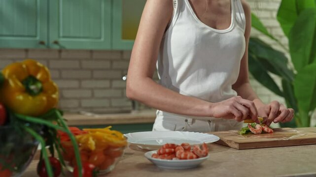 Young woman at the table in the kitchen preparing spring rolls for healthy lunch, takes thin wrapper piece, puts ingredients inside and makes a roll. Slow motion.