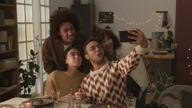 Four smiling friends taking silly selfies, gathering around food-filled table in cozy, eco-friendly room adorned with string lights