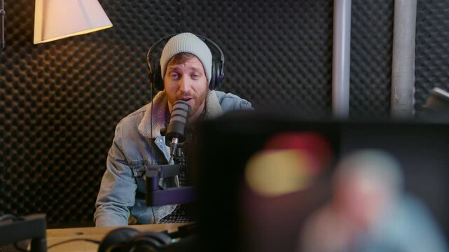 Professional podcast host or radio dj speaker talks to video camera while in sound studio. Using microphone and audio gear he films himself in sound proof booth. Excited and happy to stream live show