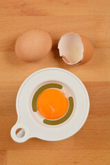 A white plastic egg separator with a bright orange yolk inside is placed next to a whole egg and a cracked eggshell on a wooden surface