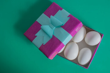 Purple gift box with blue ribbon and white eggs on a green background. Top view