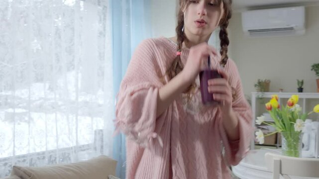 A teenage girl in a pink knitted sweater is drinking a freshly blended berry smoothie from a glass. Healthy nutrition, homemade drinks, and a balanced diet for kids and teenagers