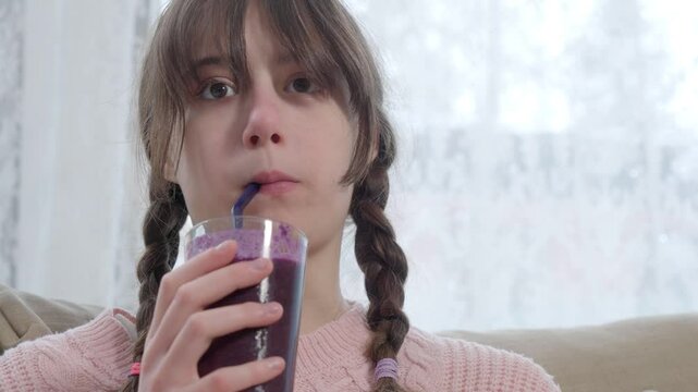 A teenage girl in a pink knitted sweater is drinking a freshly blended berry smoothie from a glass. Healthy nutrition, homemade drinks, and a balanced diet for kids and teenagers