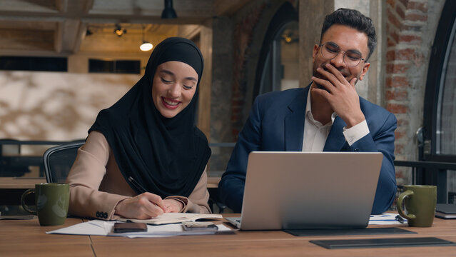 Two business colleagues co-workers talk discuss corporate work at office cafe multiracial male female employees Indian man muslim woman in hijab diverse businesswoman businessman write tasks laptop - Powered by Adobe