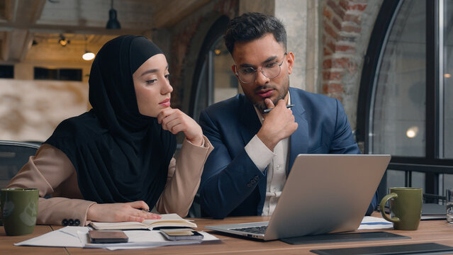Two business colleagues co-workers talk discuss corporate work at office cafe multiracial male female employees Indian man muslim woman in hijab diverse businesswoman businessman write tasks laptop