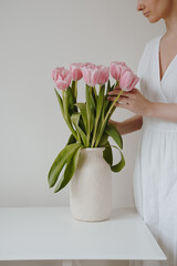Young woman touching pink tulip flowers bouquet in clay vase over white background. Minimalist floral concept