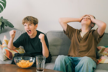 Two excited teenager boys watching match game together at home. Brothers seated on couch with...