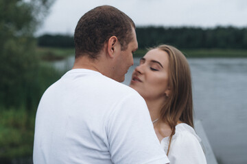 Obraz premium Romantic couple kissing and embracing near a lake and forest on a cloudy day.