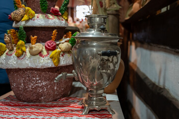 A vintage metal samovar on a traditional embroidered cloth, set in a rustic interior with decorative festive bread in the background.