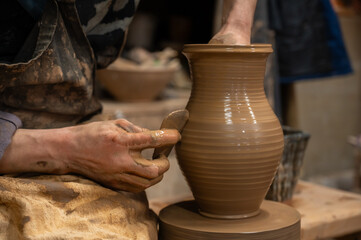 Close-up of a potter’s hands covered in wet clay as they carefully shape a ceramic vase on a...