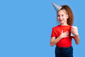 Smiling girl in red T-shirt and shiny party hat places hand on chest, looking touched and grateful....
