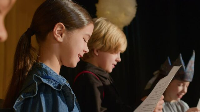 Group of diverse boys and girls holding script papers, standing on theater stage, reading out their lines