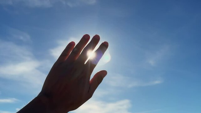 Close-up hand in sunshine outdoors, stretching arm to sun rays on blue sky with white clouds. Nature concept.