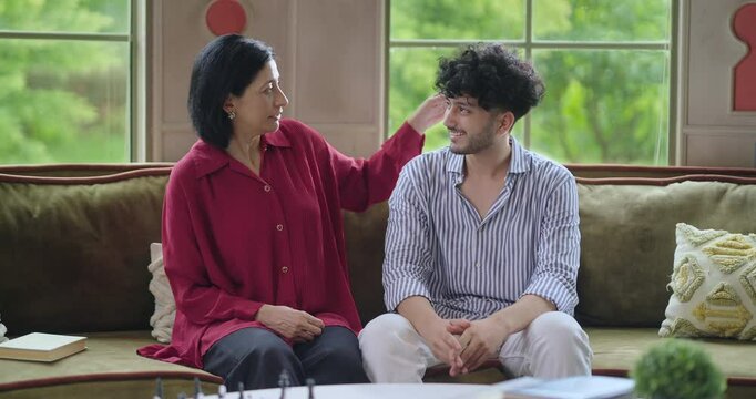 Indian happy mature woman lady with young man male sitting on cozy sofa couch talk gossip share story indoor home adult mom teen son boy speak funny joke enjoy day spend time have fun joy inside house
