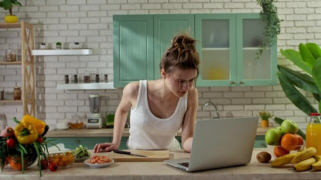 Young woman at the table in the kitchen preparing spring rolls for healthy lunch, woman cutting ingredients and watching recipe video on laptop. Slow motion.