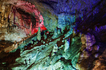 A cave with stalagmites and stalactites, illuminated with colorful lighting that highlights the natural rock formations. The textured walls and mineral structures create an ethereal atmosphere. 