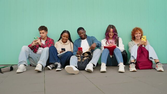 Classmate student friends relaxing together after class using mobile phone appe sitting outside. Laughing and chatting focused on their smartphones