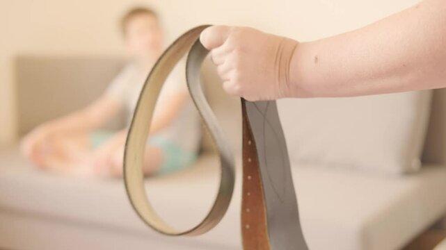 wide leather belt in female parent's hand threatens misbehaving teenage boy sitting on couch, Parenting struggles