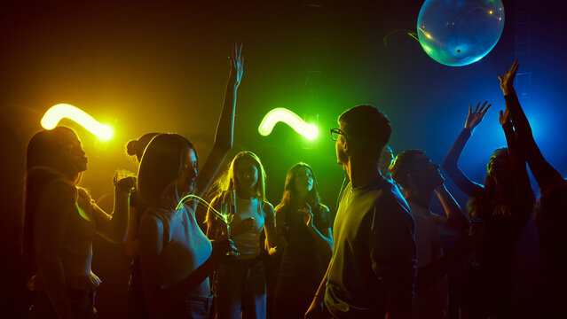 Friends gathered for party in a dimly lit club with colorful lights. Young men and women enjoying music and the youthful, carefree moments. Concept o entertainment, leisure, youth culture