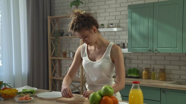 Young woman at the table in the kitchen preparing spring rolls for healthy lunch, takes thin wrapper piece, puts greens, veggies and shrimps inside and makes a roll.