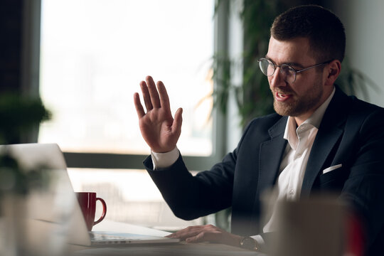 Entrepreneur in a suit having a video call, smiling and waving at his laptop. Concept of remote work, virtual meetings, online communication, professional networking, and business technology - Powered by Adobe