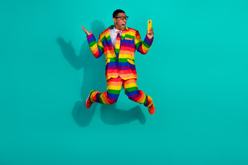 Energetic guy in a rainbow suit jumps joyfully on a teal background while holding a vibrant yellow smartphone.