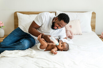Fototapeta premium Loving African father feeding his baby with a bottle while lying on a cozy bed. A heartwarming moment of fatherhood, bonding, and care, symbolizing modern parenting and nurturing relationships.