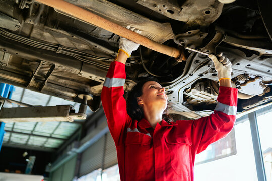 Female mechanic in red uniform working under a lifted car in an auto repair shop. Smiling while fixing the vehicle with a wrench. Concept of women in skilled trades and car maintenance.
