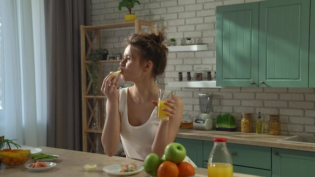Young woman at the table in the kitchen preparing spring rolls for healthy lunch, eating fresh spring rolls and drinks orange juice, enjoying the taste.