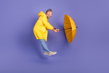 Elderly man in vibrant yellow raincoat holding bright umbrella against violet background,...