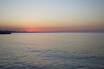Sunset by the sea, purple sky, houses in the distance, sunset with orange-purple sky, sunset reflected gentle in the sea