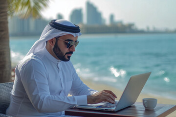 An Arab man wearing ethnic clothes sits at a beachfront cafe, working on his laptop. City scape, natural sea view. Modern business work scene shot.