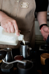 barista in apron pours milk into metal frothing pitcher, preparing coffee drink. scene captures focus and skill involved in crafting perfect beverage, with various coffee making tools visible