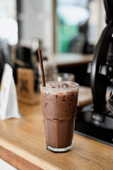 tall glass of iced chocolate drink with straw sits on wooden counter in cozy cafe setting, surrounded by blurred background elements