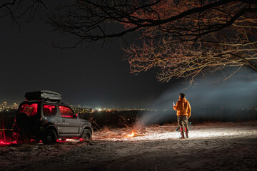 A man stands with a lantern on his head. lights of the night city on background. A bonfire burns with smoke. An SUV vehicle is parked on top. The concept of recreation and trips to nature.