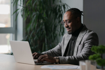 Professional businessman working on a laptop in a modern office, wearing a stylish suit and glasses. Concept of productivity, remote work, corporate lifestyle, and business success