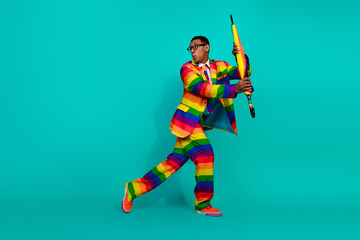 Obraz premium Young man in vibrant rainbow suit holding colorful umbrella on turquoise background, expressing joy and individuality