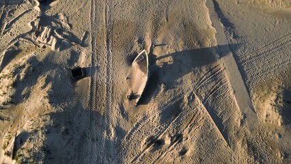 Aerial view of an abandoned boat partially buried in sand, surrounded by tire tracks and...