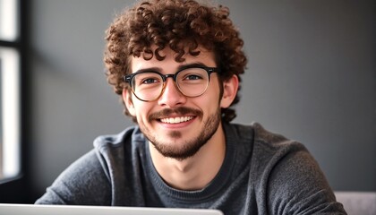 Fototapeta premium A young man with curly hair and glasses is smiling at the camera while sitting behind a laptop.
