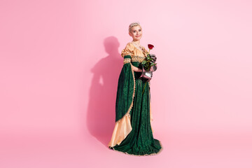 Fototapeta premium Elegant young woman in a historical green dress holding a rose on a pink background