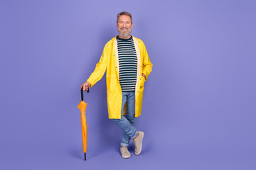 Smiling mature man in a yellow raincoat holding an umbrella against a purple background, dressed in a casual striped shirt