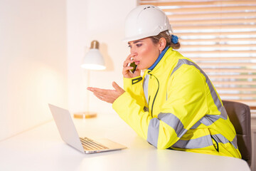 Portrait of female Construction worker in safety gear communicates with a client at a desk while using a laptop in a well-lit office environment