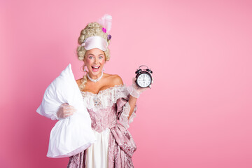Elegant woman in vintage dress holding pillow and clock against pink backdrop, depicting humor and...