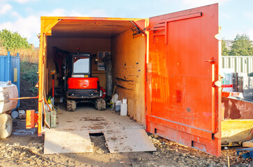 Construction site storage container houses a compact excavator ready for use on a sunny day at a work site