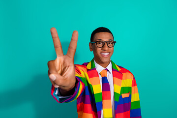 Young man in a vibrant rainbow-patterned suit against a teal background showing a peace gesture and smiling