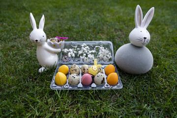 Two souvenir porcelain Easter bunny on a green lawn next to quail and multi-colored chocolate eggs. Home decor, festive Easter