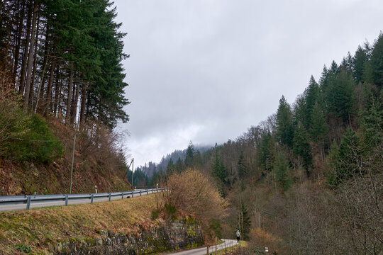 Scenic Mountain Road with Traditional Houses and Red Roofs in Black Forest Valley Landscape. Winding Rural Highway Through Alpine Village with White Buildings and Green Forest Hills Under Cloudy Sky.