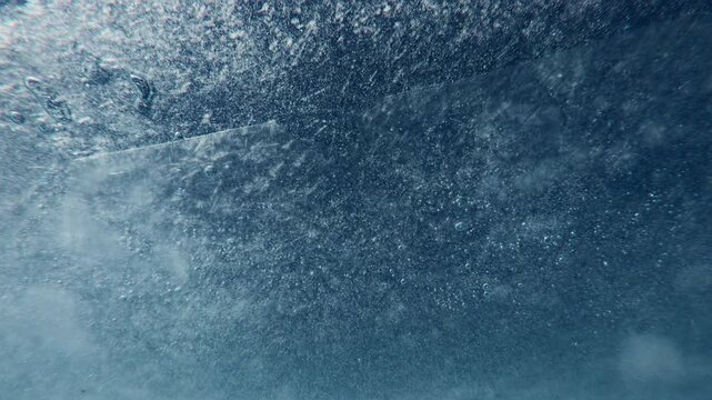 A close-up of the thin spring melt ice moving along the water current. Underwater view of ice texture with frozen air bubbles against blue sky.