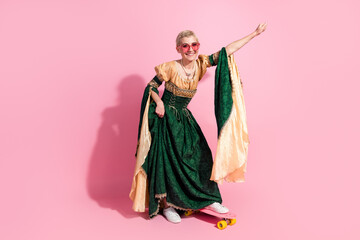 Fototapeta premium Young woman in elegant green dress skateboarding on pink background showcasing medieval attire with modern twist